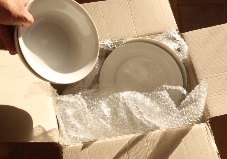 Cardboard Packing Box, Plates and Bubble Wrap. Cardboard packing box open on wooden floor with bubble wrap inside. Bowls being placed inside with plates.